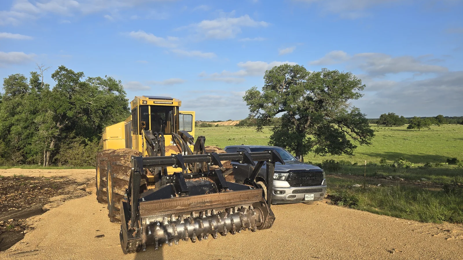 Wheeled forestry mulcher and a pickup on a caliche pad beside a green pasture, forestry mulching