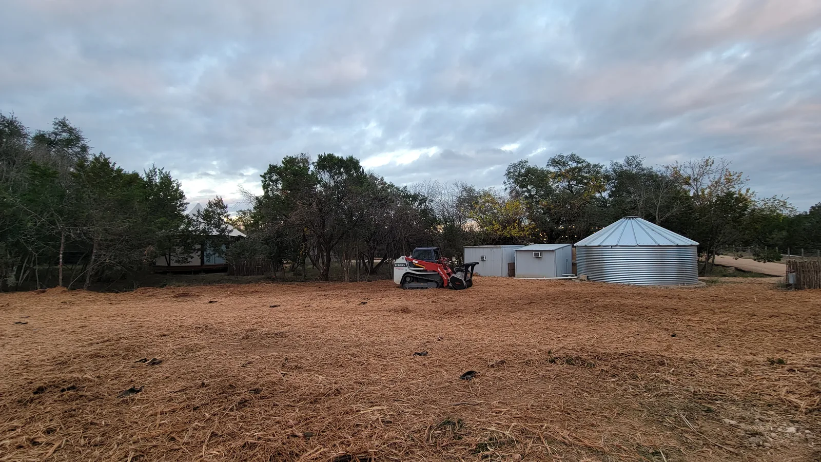 Freshly mulched field with a tracked skid steer mulcher near the sheds and a water tank, land clearing