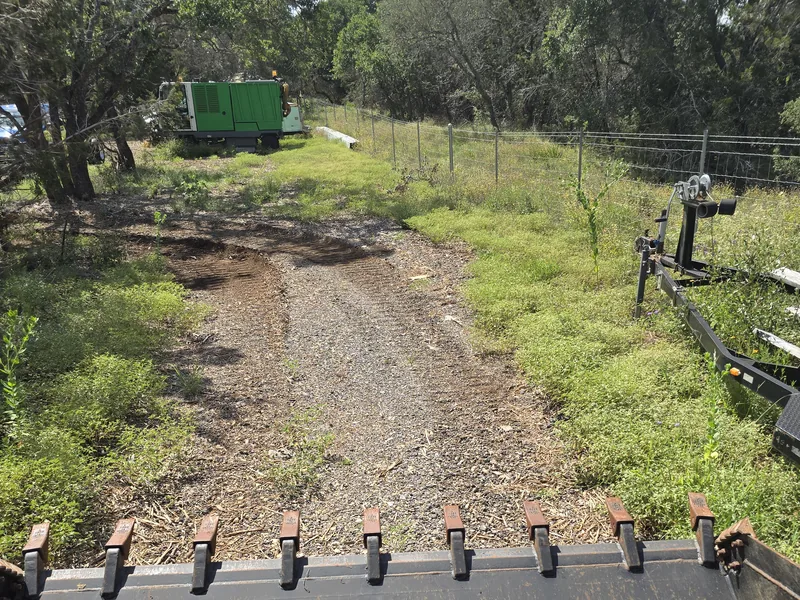 Cleared gravel strip running beside a wire fence, seen over a skid steer bucket, fence line clearing