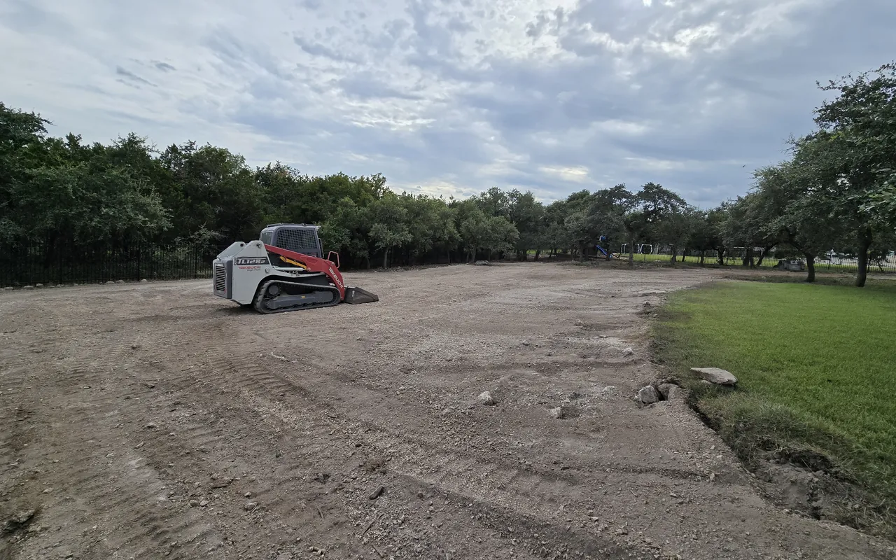 Tracked skid steer parked on freshly graded, level ground beside a green lawn and oaks, land grading