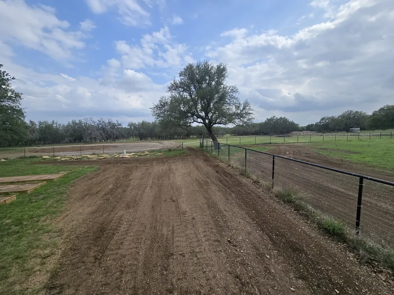 Freshly graded strip of dirt along a black pipe fence, with an oak and pasture beyond, land grading