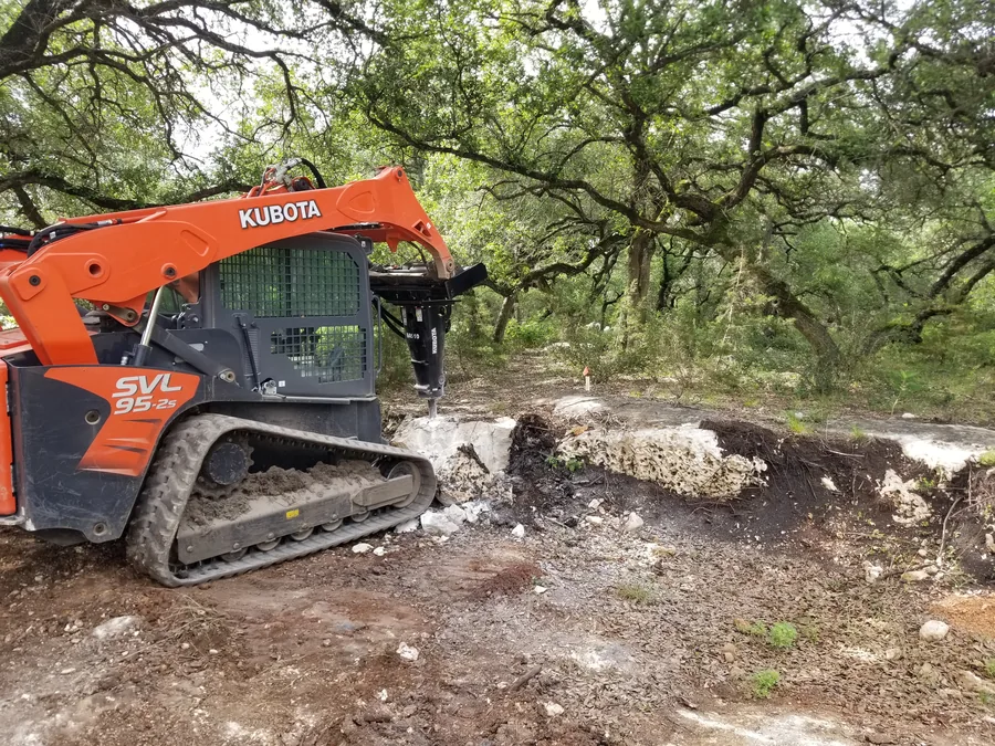 Tracked skid steer with a hydraulic hammer breaking a limestone ledge under oaks, light demolition